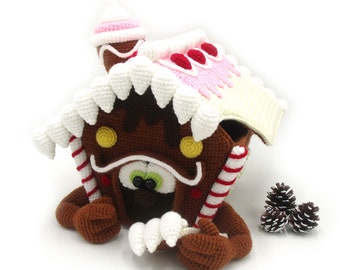 May include: A crocheted gingerbread house toy with candy cane accents and a brown bear face. The house is decorated with white frosting, red candy hearts, and yellow candy dots.