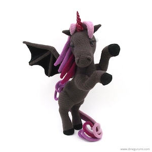 May include: A crocheted plush toy unicorn with a dark grey body, pink and purple mane and tail, and black bat wings.