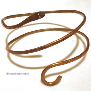May include: A copper-colored arm cuff in a spiral design. The cuff wraps around the arm with a snake-like appearance. The cuff has a hammered texture and a hook-and-eye closure. The text "@mixedmythologies" is in the lower left corner.