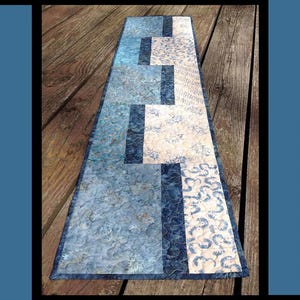 May include: A quilted table runner with a blue and white floral pattern. The runner features a rectangular design with the text "Ripple Runner". The design includes various shades of blue and white.