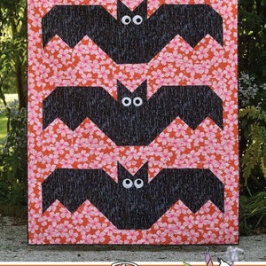 Puede incluir: Un edredón con temática de Halloween con tres apliques de murciélagos negros con ojos blancos sobre un fondo floral rosa. La palabra "Batty" está en la parte superior, con "Running Doe Quilts" y un logotipo en la parte inferior. Un artículo festivo hecho a mano.