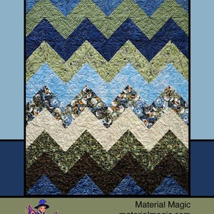 May include: A quilted wall hanging with a mountain design. The quilt features a zig-zag pattern in shades of blue, green, and cream, resembling mountain peaks. The word "Mountains" is written in a script font at the top. The quilt is framed in a black border.
