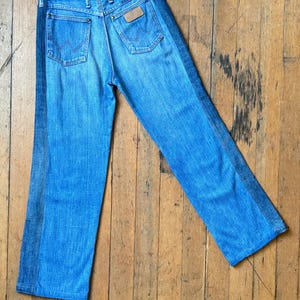Pu&ograve; includere: Jeans di denim blu chiaro con una striscia blu scuro a contrasto sul lato di ogni gamba. I jeans hanno due tasche posteriori e una toppa in pelle marrone con un logo. I jeans sono appoggiati su una superficie di legno.