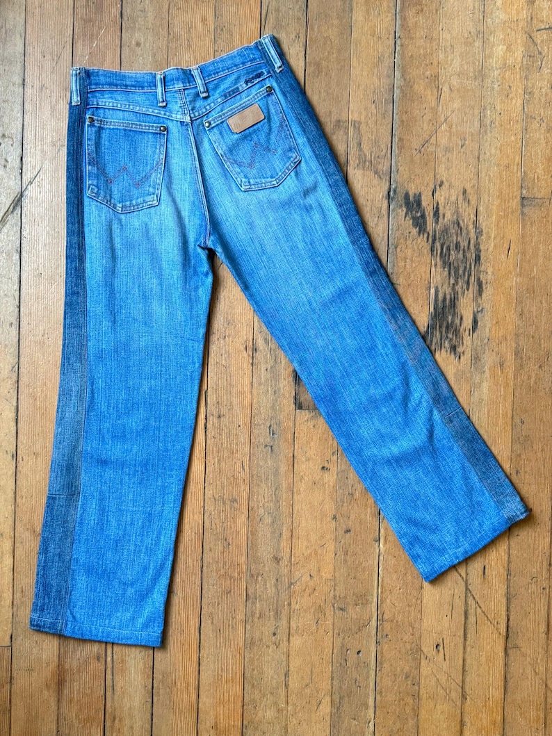 Pu&ograve; includere: Jeans di denim blu chiaro con una striscia blu scuro a contrasto sul lato di ogni gamba. I jeans hanno due tasche posteriori e una toppa in pelle marrone con un logo. I jeans sono appoggiati su una superficie di legno.
