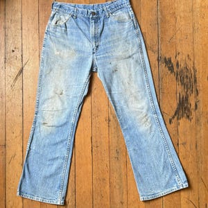 May include: Light blue denim jeans with a faded appearance. The jeans have a classic five-pocket design, a button and zipper closure, and a flared leg. The fabric shows signs of wear and tear, adding to their vintage appeal.
