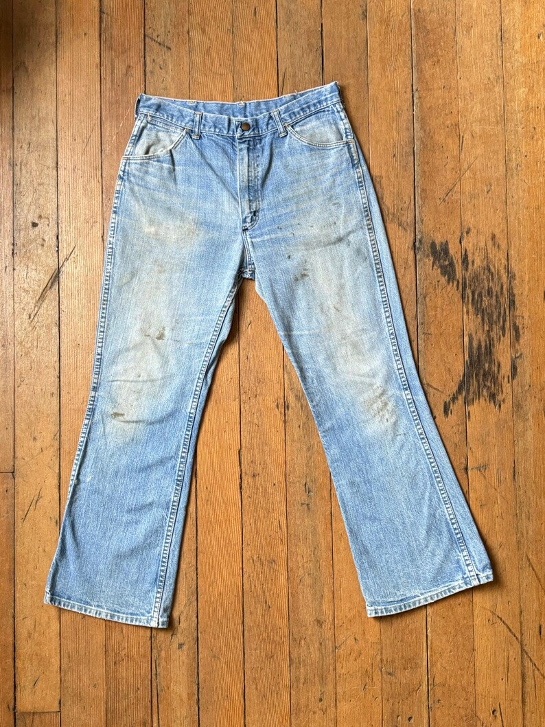 May include: Light blue denim jeans with a faded appearance. The jeans have a classic five-pocket design, a button and zipper closure, and a flared leg. The fabric shows signs of wear and tear, adding to their vintage appeal.