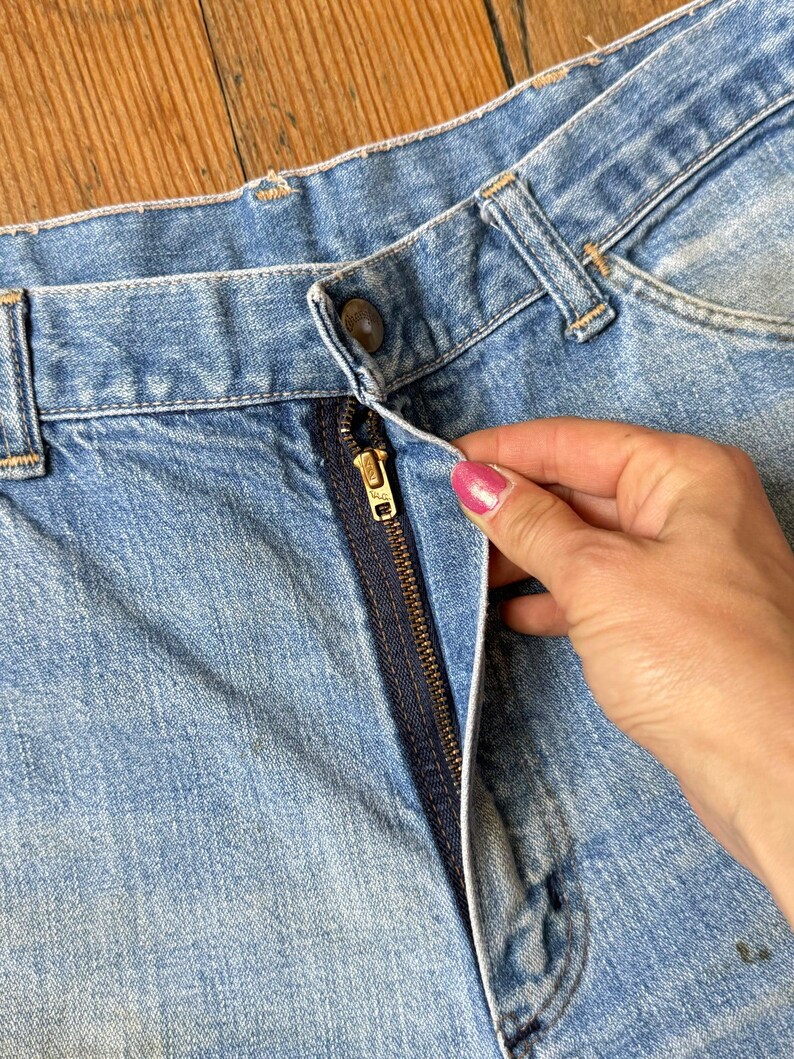 May include: Close-up of light blue denim jeans with a visible zipper and button closure. The jeans show signs of wear and tear, with a slightly faded appearance. The zipper is a dark navy blue color.