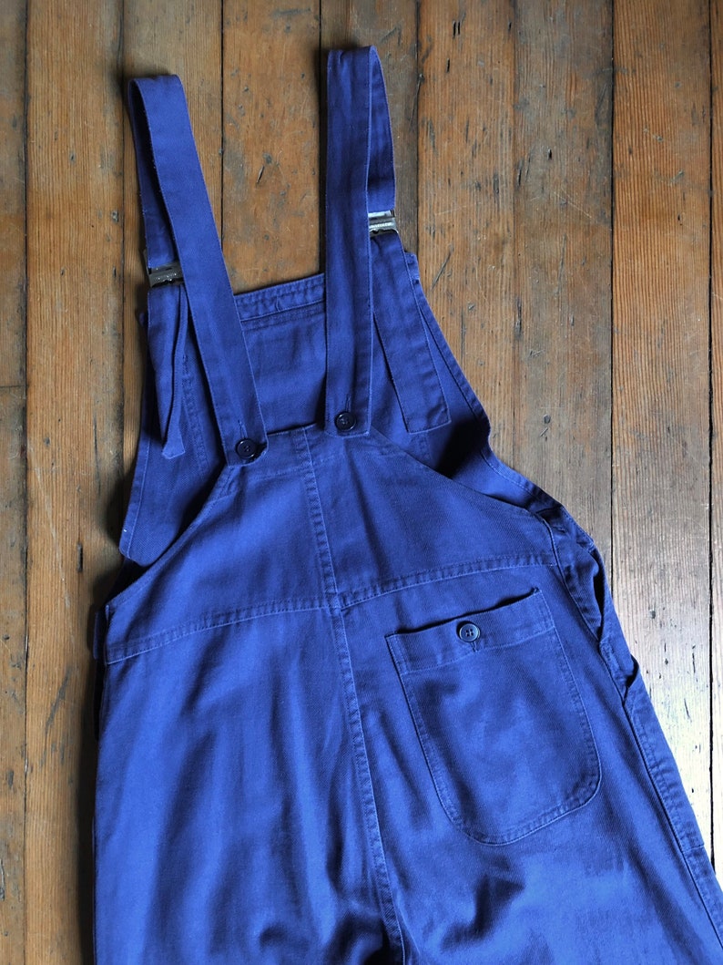 K&ouml;nnte beinhalten: Eine blaue Denim-Latzhose mit verstellbaren Tr&auml;gern und einer einzigen Ges&auml;&szlig;tasche.