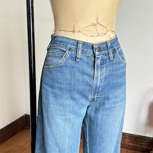 Pu&ograve; includere: Jeans di denim blu chiaro con un classico design a cinque tasche. I jeans hanno una chiusura con bottone e cerniera, passanti per cintura e una vestibilit&agrave; a gamba dritta. I jeans sono esposti su un manichino.