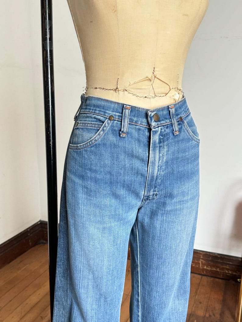 Pu&ograve; includere: Jeans di denim blu chiaro con un classico design a cinque tasche. I jeans hanno una chiusura con bottone e cerniera, passanti per cintura e una vestibilit&agrave; a gamba dritta. I jeans sono esposti su un manichino.