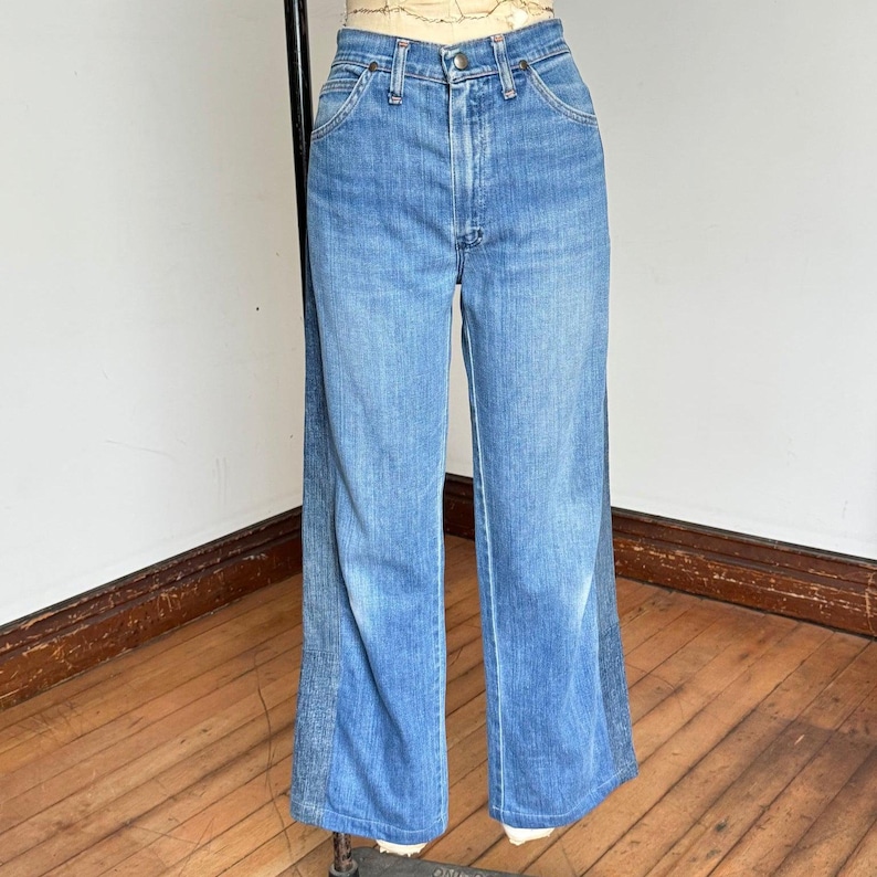 Pu&ograve; includere: Jeans di denim blu chiaro a vita alta e gamba larga. I jeans hanno un classico stile a cinque tasche e una chiusura con bottone e cerniera. Il denim ha un lavaggio sbiadito e un leggero svasatura sull'orlo.