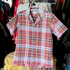 Pode incluir: Camisa de manga curta com estampa xadrez vermelha, preta, amarela e azul clara. A camisa tem gola de acampamento e um bolso no peito. A camisa vintage está pendurada em um cabide.