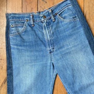 Pu&ograve; includere: Jeans di denim blu chiaro con una striscia verticale blu scuro sul lato. I jeans hanno un classico design a cinque tasche, una chiusura con bottone e cerniera e rivetti color rame. Il denim ha un aspetto sbiadito e usurato.