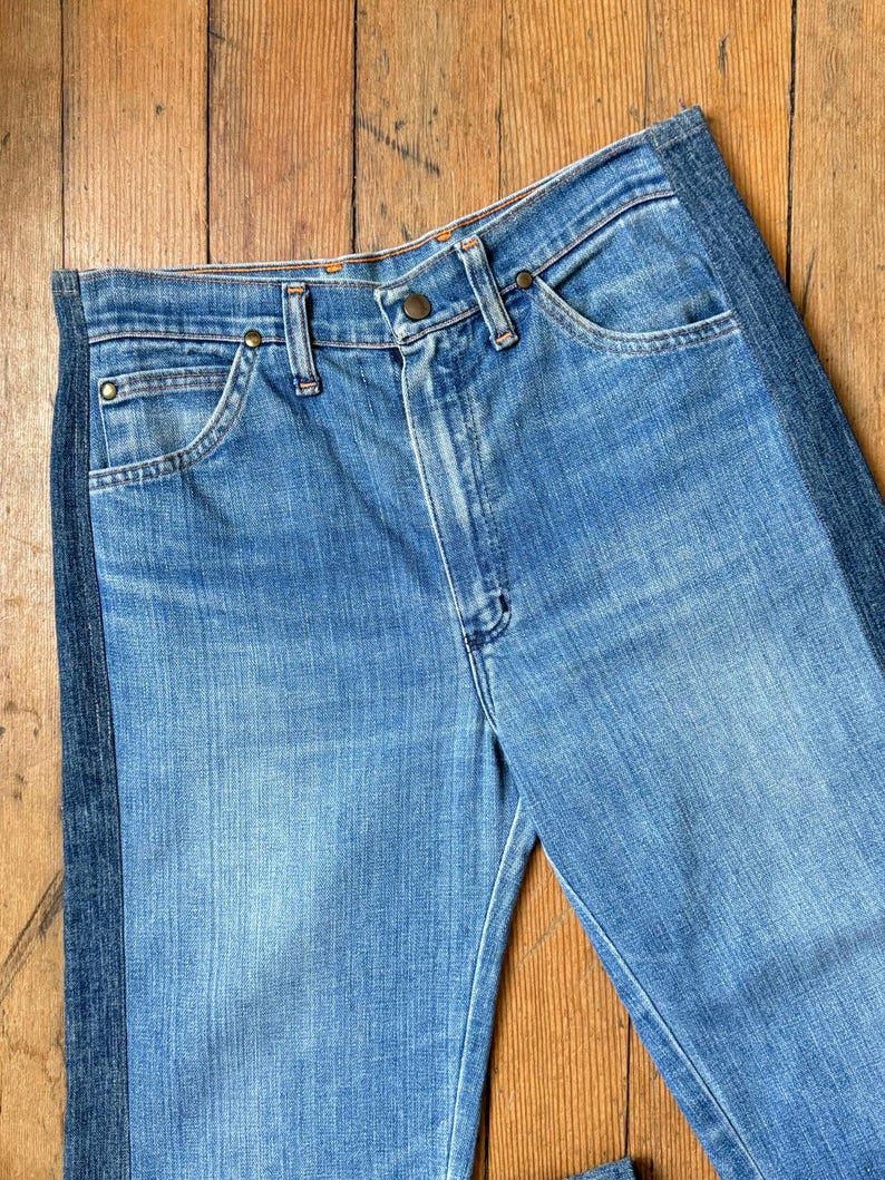 Pu&ograve; includere: Jeans di denim blu chiaro con una striscia verticale blu scuro sul lato. I jeans hanno un classico design a cinque tasche, una chiusura con bottone e cerniera e rivetti color rame. Il denim ha un aspetto sbiadito e usurato.