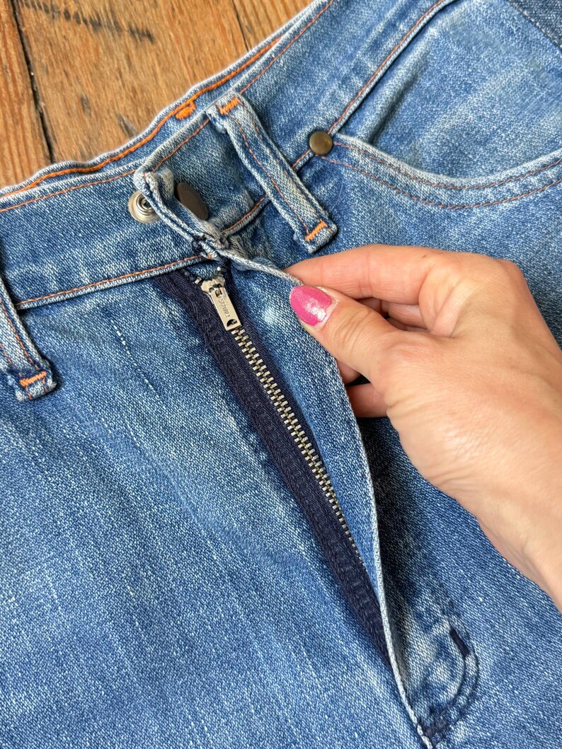 Pu&ograve; includere: Primo piano di jeans blu denim con chiusura a cerniera e bottone. I jeans hanno cuciture arancioni e una cerniera blu scuro. La cerniera &egrave; parzialmente aperta.