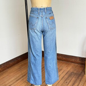 Pu&ograve; includere: Jeans Wrangler blu chiaro in denim con vita alta e gambe svasate. I jeans hanno il classico stile a cinque tasche, una toppa in pelle marrone e il logo Wrangler. I jeans sono esposti su un manichino.