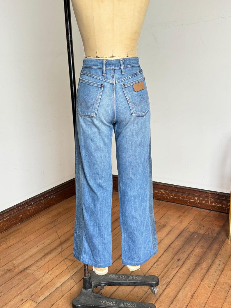 Pu&ograve; includere: Jeans Wrangler blu chiaro in denim con vita alta e gambe svasate. I jeans hanno il classico stile a cinque tasche, una toppa in pelle marrone e il logo Wrangler. I jeans sono esposti su un manichino.