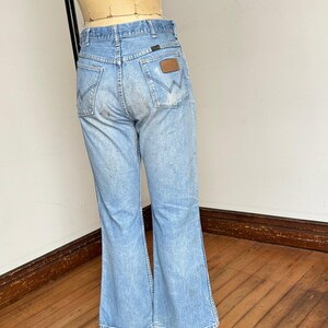 May include: Light blue denim jeans with a flared leg, displayed on a mannequin. The jeans feature classic five-pocket styling, a brown leather patch on the back pocket, and the Wrangler logo.