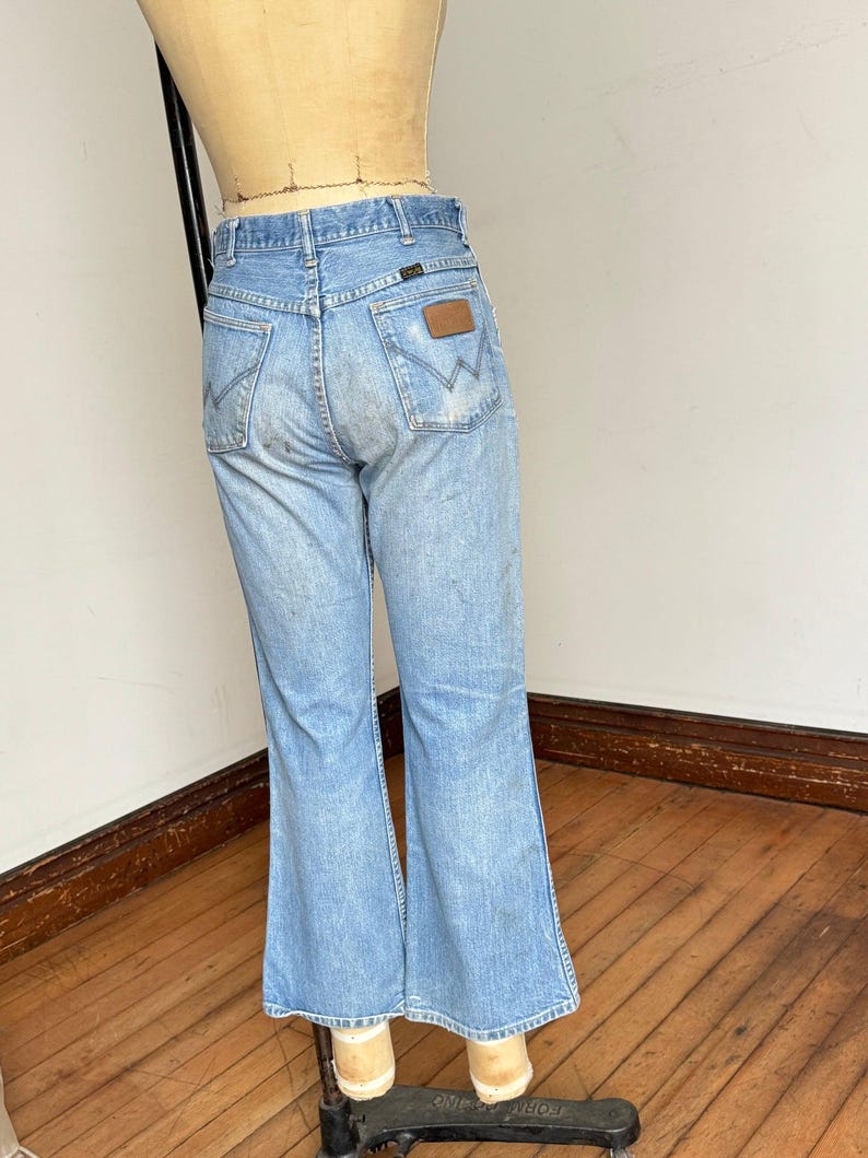 May include: Light blue denim jeans with a flared leg, displayed on a mannequin. The jeans feature classic five-pocket styling, a brown leather patch on the back pocket, and the Wrangler logo.