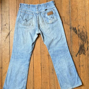 May include: Light blue denim jeans with a classic five-pocket design. The jeans feature a faded wash and a brown leather patch on the back pocket. The jeans are laid flat on a wooden surface.