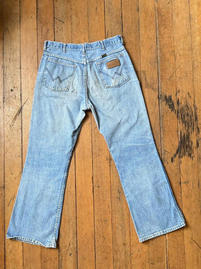May include: Light blue denim jeans with a classic five-pocket design. The jeans feature a faded wash and a brown leather patch on the back pocket. The jeans are laid flat on a wooden surface.
