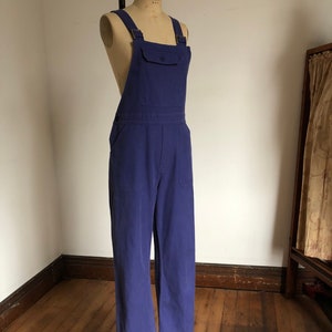K&ouml;nnte beinhalten: Eine blaue Denim-Latzhose mit einer einzigen Tasche vorne. Die Latzhose befindet sich auf einem Mannequin und hat verstellbare Tr&auml;ger.