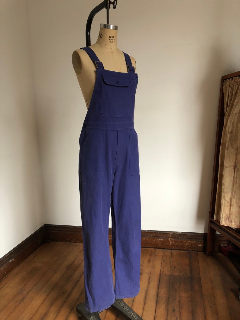 K&ouml;nnte beinhalten: Eine blaue Denim-Latzhose mit einer einzigen Tasche vorne. Die Latzhose befindet sich auf einem Mannequin und hat verstellbare Tr&auml;ger.