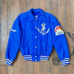 Può includere: Giacca da softball vintage blu reale con dettagli bianchi. La giacca presenta la scritta "Manager" sul petto sinistro e "Lahr Ladies Softball 1979" sul petto destro. La manica destra ha una toppa con il nome "Marie".