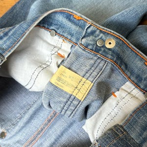 Pu&ograve; includere: Primo piano di jeans in denim blu con cuciture arancioni ed etichetta gialla. I jeans hanno un classico design a cinque tasche e una chiusura con bottone e cerniera. L'etichetta recita "Wrangler".