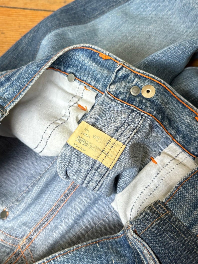 Pu&ograve; includere: Primo piano di jeans in denim blu con cuciture arancioni ed etichetta gialla. I jeans hanno un classico design a cinque tasche e una chiusura con bottone e cerniera. L'etichetta recita "Wrangler".