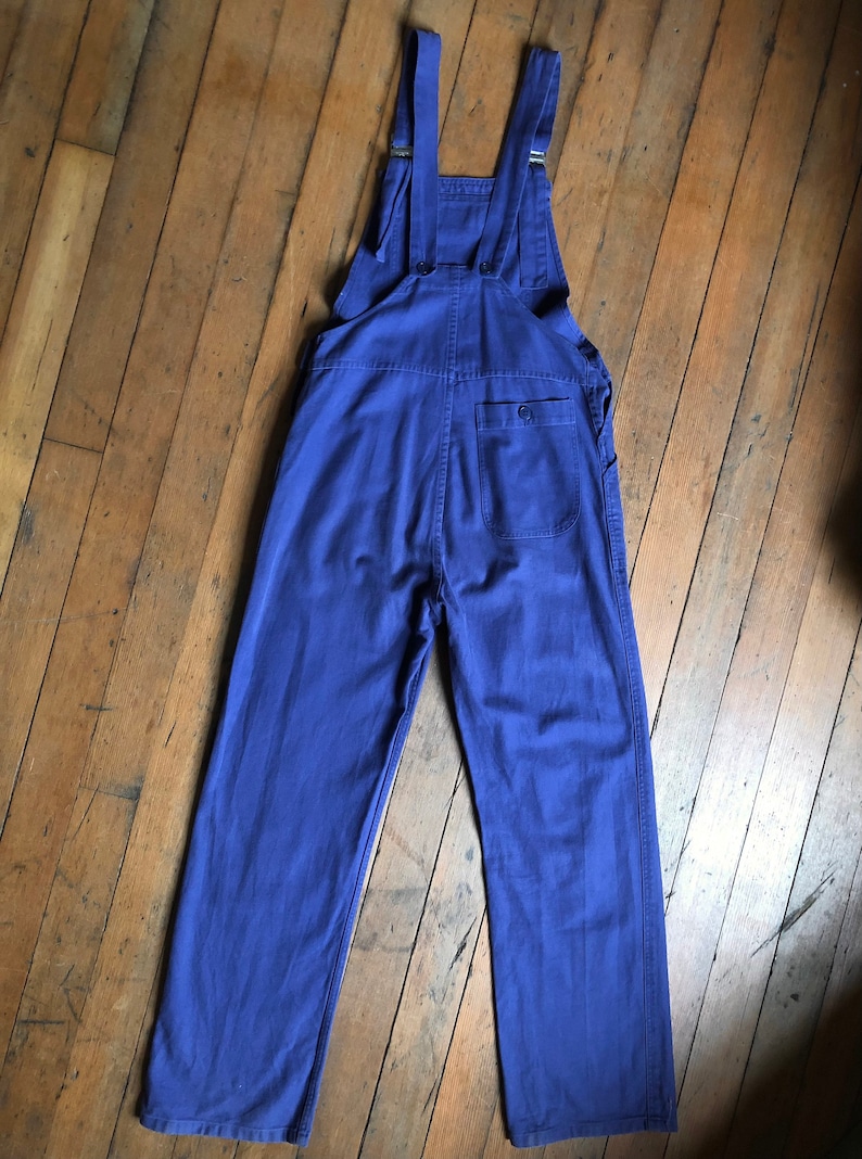 K&ouml;nnte beinhalten: Eine blaue Denim-Latzhose mit verstellbaren Tr&auml;gern und einer einzigen Ges&auml;&szlig;tasche.