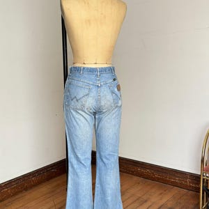 May include: Light blue denim jeans with a flared leg design. The jeans are displayed on a mannequin, showcasing the back pockets and the Wrangler logo. The jeans have a classic, vintage look.