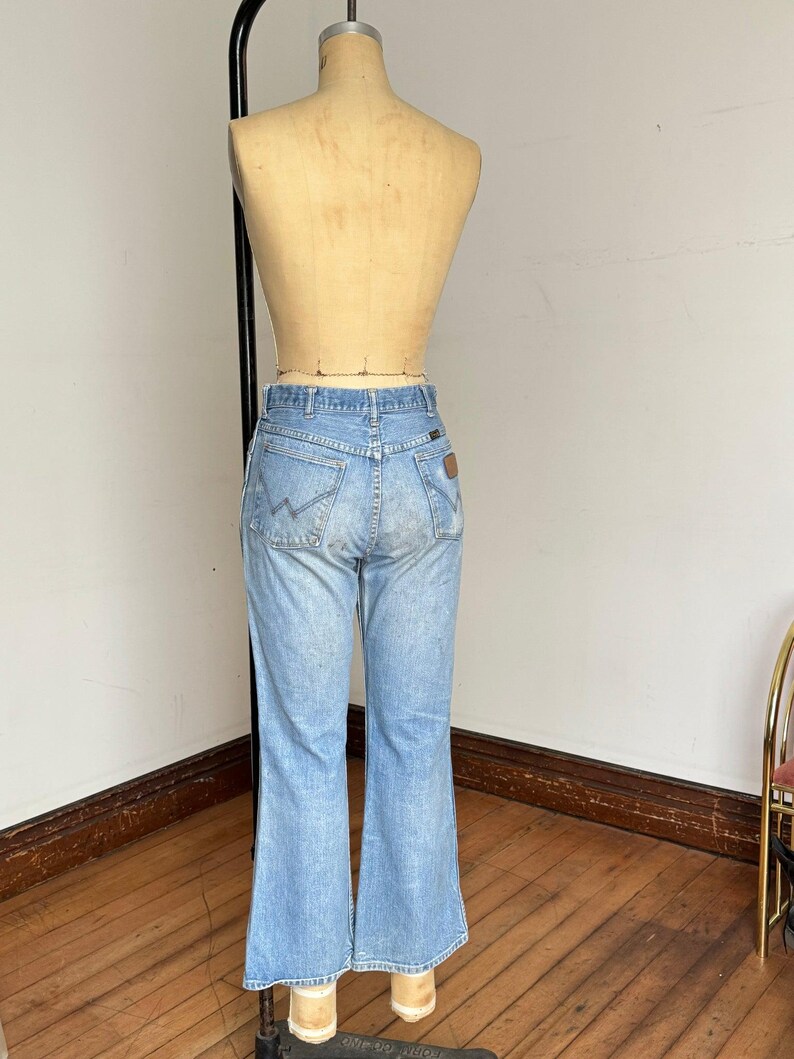 May include: Light blue denim jeans with a flared leg design. The jeans are displayed on a mannequin, showcasing the back pockets and the Wrangler logo. The jeans have a classic, vintage look.