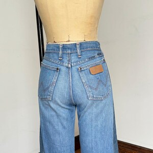 Pu&ograve; includere: Jeans in denim blu chiaro con vita alta e gamba larga. I jeans presentano due tasche posteriori con cuciture decorative e una toppa in pelle marrone. I jeans sono esposti su un manichino.