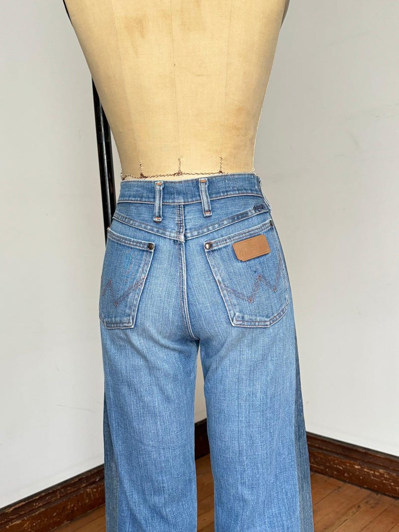 Pu&ograve; includere: Jeans in denim blu chiaro con vita alta e gamba larga. I jeans presentano due tasche posteriori con cuciture decorative e una toppa in pelle marrone. I jeans sono esposti su un manichino.