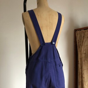 K&ouml;nnte beinhalten: Eine blaue Denim-Latzhose mit verstellbaren Tr&auml;gern und einem einzigen Knopfverschluss auf der R&uuml;ckseite. Die Latzhose hat eine aufgesetzte Tasche auf der rechten Hinterseite.