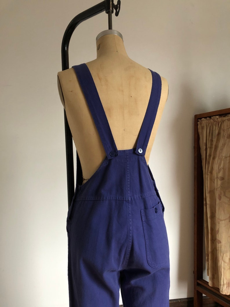 K&ouml;nnte beinhalten: Eine blaue Denim-Latzhose mit verstellbaren Tr&auml;gern und einem einzigen Knopfverschluss auf der R&uuml;ckseite. Die Latzhose hat eine aufgesetzte Tasche auf der rechten Hinterseite.