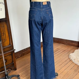 Puede incluir: Vaqueros acampanados de mezclilla azul oscuro. Los vaqueros tienen una cintura alta y presentan la etiqueta de la marca Levi's clásica en la cinturilla trasera. Los bolsillos traseros tienen las costuras características de Levi's. Los vaqueros se exhiben en un maniquí.