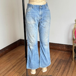 May include: Light blue denim jeans with a faded wash and slight flare at the hem. The jeans show signs of wear with some discoloration and stains. The jeans are displayed on a mannequin.