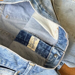 May include: Close-up of light blue denim jeans. The jeans have a button closure and a label that reads "Wrangler". The jeans are size 16 Husky. The denim has a worn appearance.