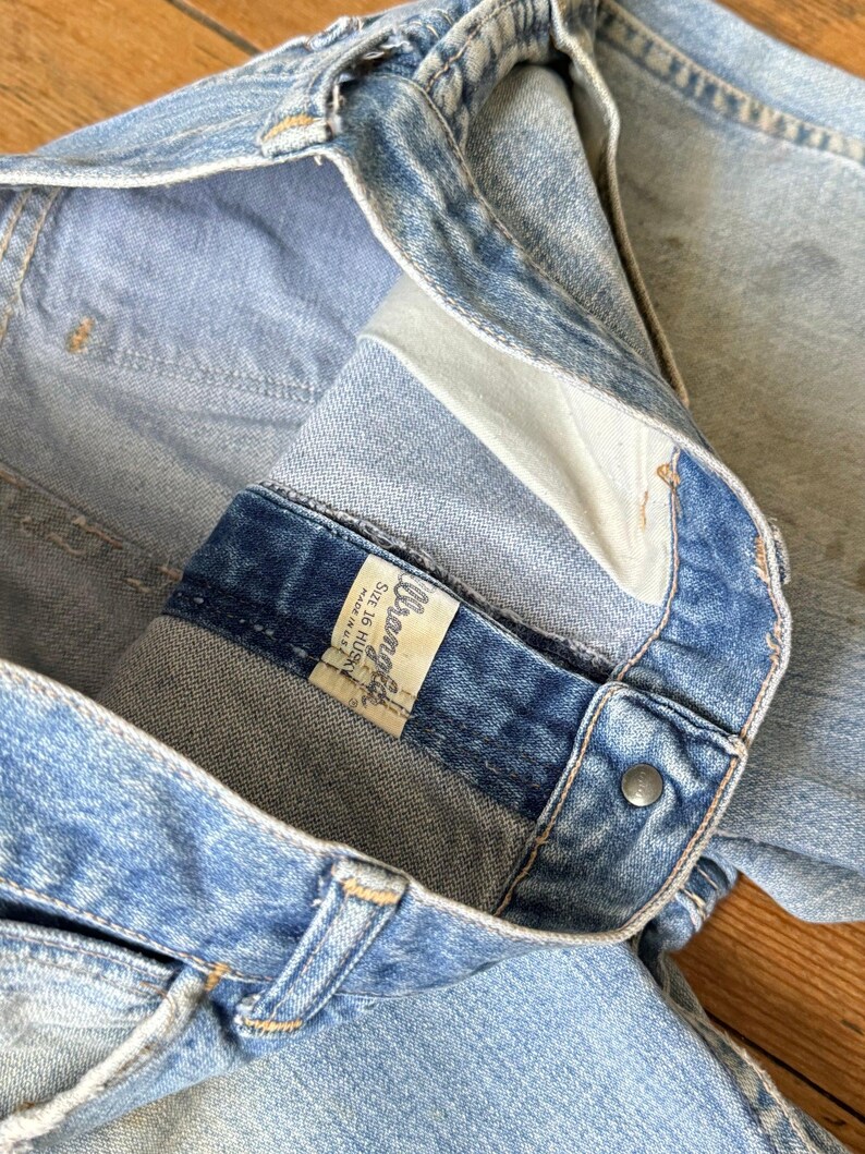 May include: Close-up of light blue denim jeans. The jeans have a button closure and a label that reads "Wrangler". The jeans are size 16 Husky. The denim has a worn appearance.