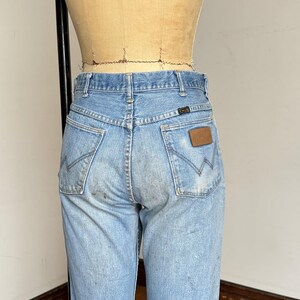 May include: Light blue denim jeans with a classic fit, featuring the Wrangler logo on the back pocket. The jeans show signs of wear and tear, adding to their vintage appeal. The back pockets have the signature Wrangler stitching.