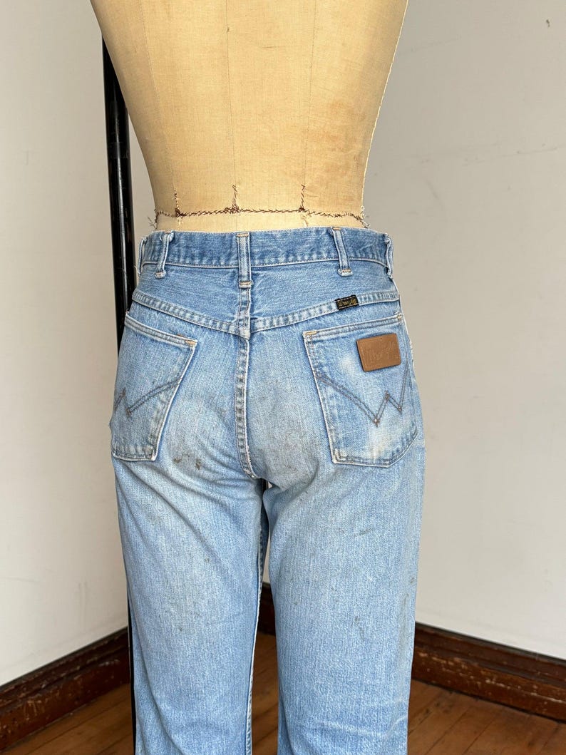 May include: Light blue denim jeans with a classic fit, featuring the Wrangler logo on the back pocket. The jeans show signs of wear and tear, adding to their vintage appeal. The back pockets have the signature Wrangler stitching.