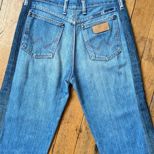 Pu&ograve; includere: Jeans in denim blu chiaro con una striscia blu scuro sul lato. I jeans hanno due tasche posteriori con un design cucito in rosso e una toppa in pelle marrone. Il marchio "Wrangler" &egrave; visibile.