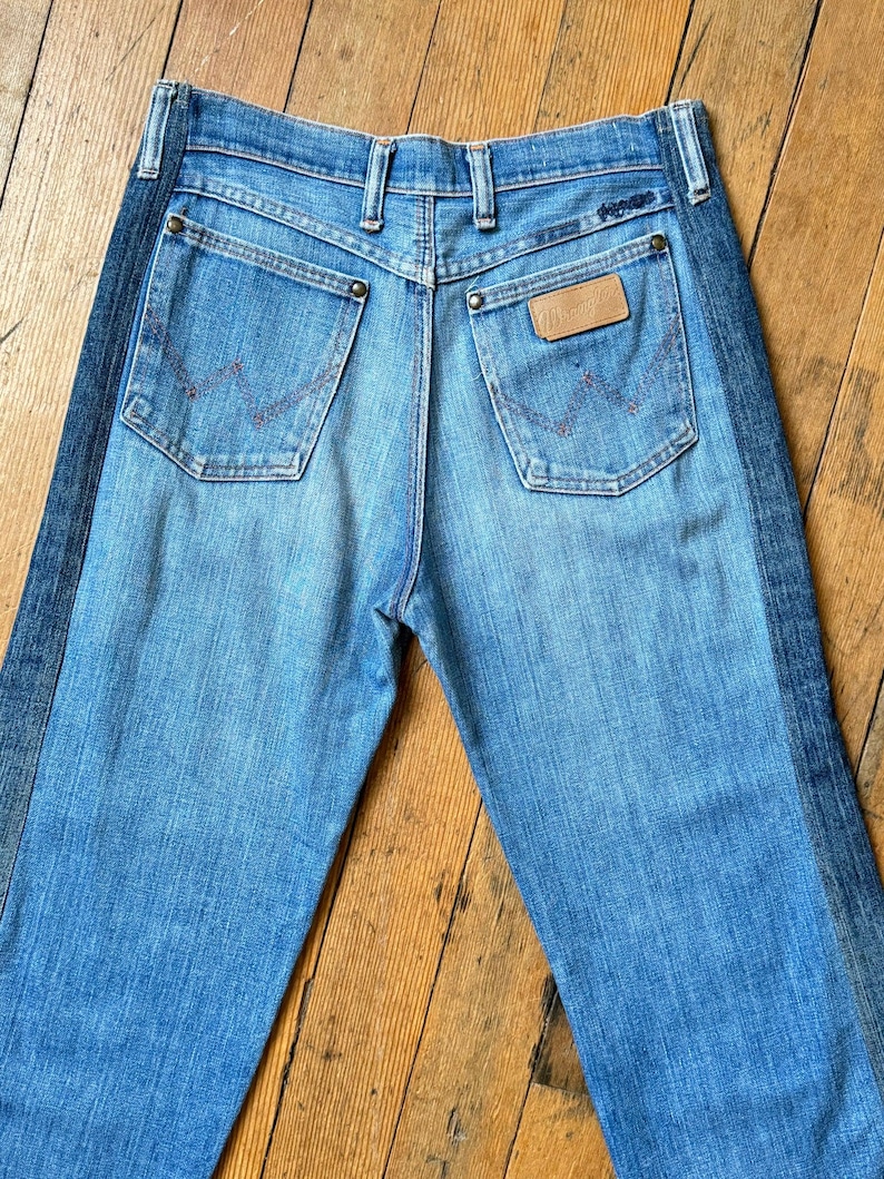 Pu&ograve; includere: Jeans in denim blu chiaro con una striscia blu scuro sul lato. I jeans hanno due tasche posteriori con un design cucito in rosso e una toppa in pelle marrone. Il marchio "Wrangler" &egrave; visibile.
