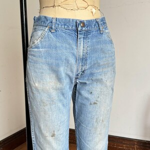 May include: Light blue denim jeans with a button closure and pockets. The jeans show signs of wear with visible stains and fading, suggesting a vintage or workwear aesthetic. The jeans are displayed on a mannequin.