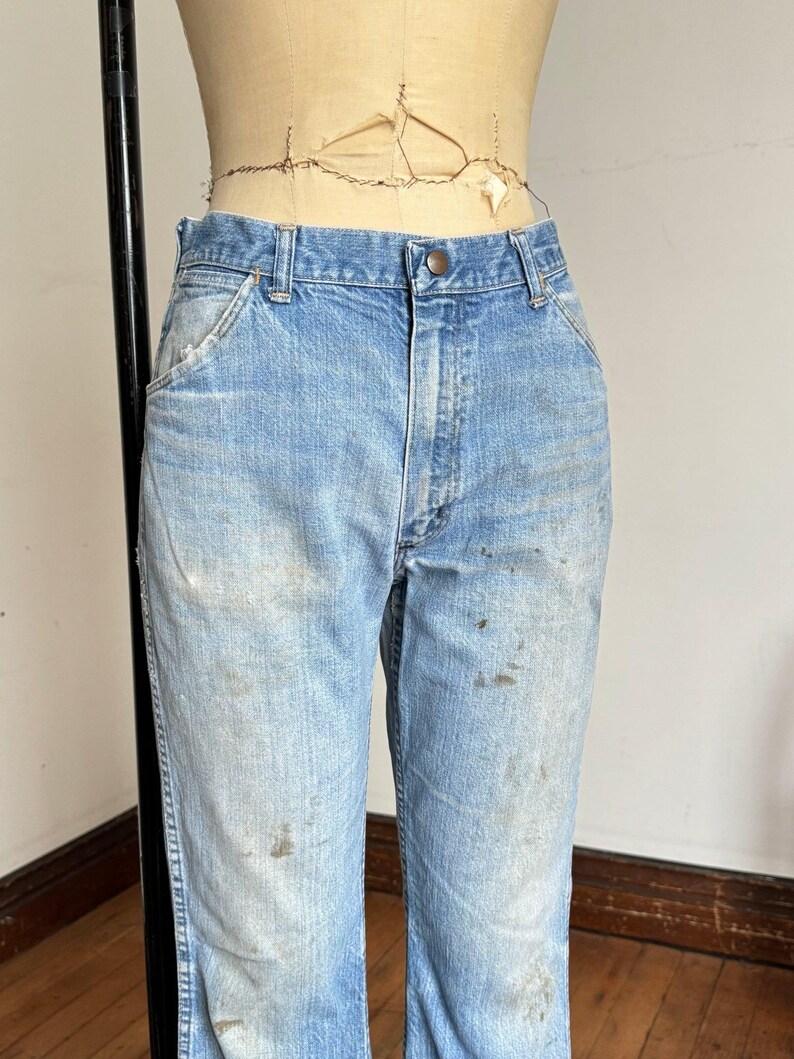 May include: Light blue denim jeans with a button closure and pockets. The jeans show signs of wear with visible stains and fading, suggesting a vintage or workwear aesthetic. The jeans are displayed on a mannequin.
