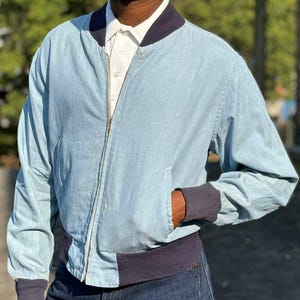 May include: A light blue bomber jacket with a navy blue ribbed collar, cuffs, and waistband. The jacket has a zip closure and a pocket on the left side.