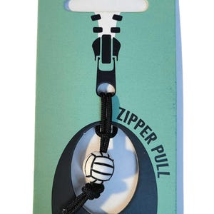 May include: A black zipper pull with a white volleyball charm. The charm is attached to a black cord. The packaging is green with a black zipper design and the text "ZIPPER PULL" and "VIDA VIBE".