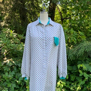 May include: A white long-sleeved shirt with a teal collar and a teal pocket on the left side. The shirt has a small, white and purple floral pattern.